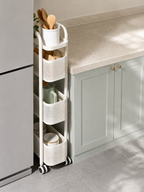 A white four-tier rolling kitchen organizer cart with woven baskets and a utensil holder, shown tucked between a refrigerator and a kitchen counter.