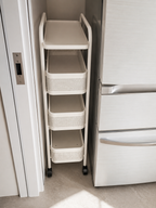 A 4-tier white rolling utility cart with perforated baskets, shown tucked between a white refrigerator and a door in a kitchen setting.