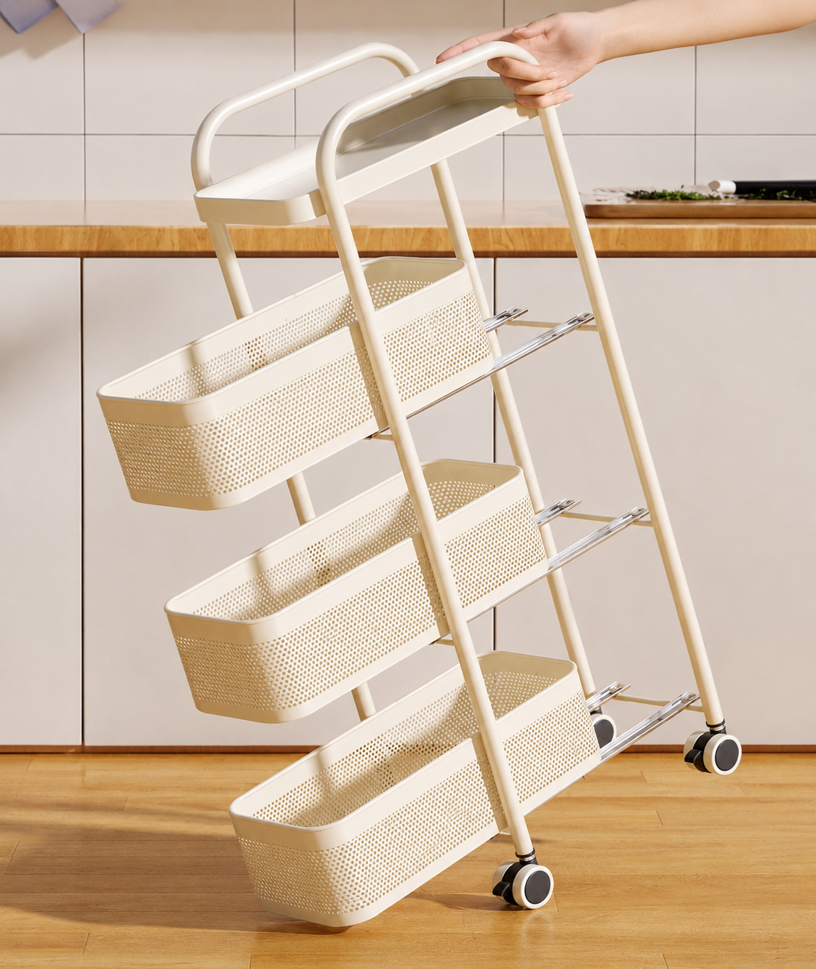 A cream-colored three-tier rolling storage cart with woven baskets and metal support bars, shown with a hand pushing its handle in a kitchen setting.