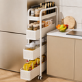 A white three-tier rolling kitchen organizer cart with woven baskets, shown tucked between a beige refrigerator and kitchen counter. The cart is stocked with spices, jars, and bottles, and a fruit bowl sits on the counter beside it.