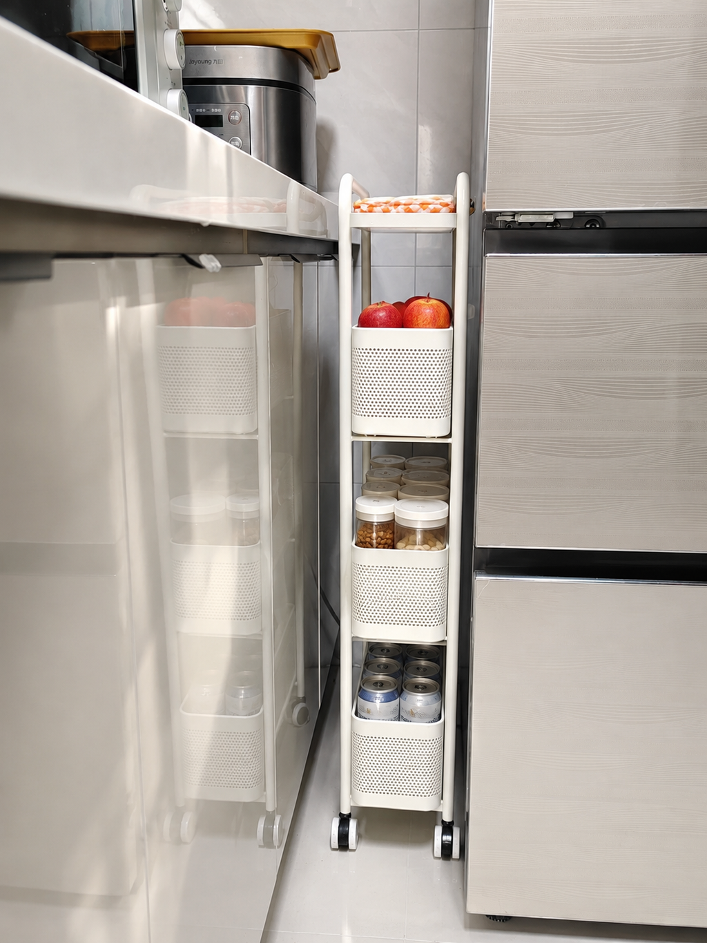 A white 4-tier rolling kitchen organizer cart with mesh baskets, shown tucked between a white countertop and a stainless steel refrigerator in a kitchen setting.